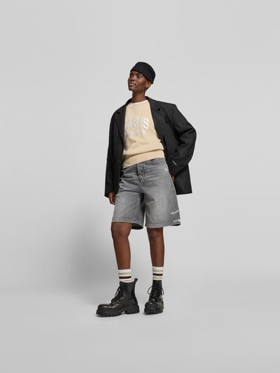 Frauenmodel trägt Sweatshirt aus reiner Baumwolle von Anine Bing in Beige mit Label-Detail, lockerer Passform und Rundhalsausschnitt, elastische Abschlüsse, kombiniert mit grauen Shorts, Ganzkörperansicht von vorne.