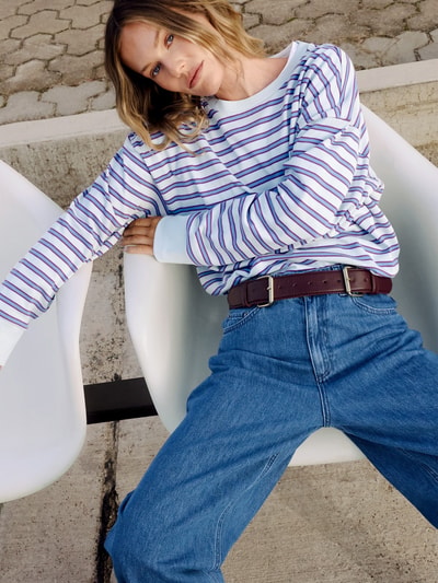 Frauenmodel trägt Longsleeve mit Streifenmuster - curated by Tinvcb von Tinvcb, weiß mit lila Streifen, lockerer Schnitt, Baumwolle, gerippte Abschlüsse, Rundhalsausschnitt, Kombination mit blauer Jeans, Nahaufnahme Oberkörper.