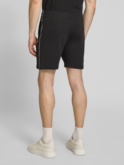 Männermodel trägt Regular Fit Sweatshorts aus reiner Baumwolle von Lacoste in Schwarz mit Logostreifen und Label Patch, Ganzkörperansicht Rückansicht.