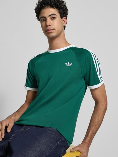 Männermodel trägt T-Shirt mit Label-Stitching von adidas Originals, bawełniany miks, krój regular fit, zielony z białymi elementami, widok na górną część ciała z przodu
