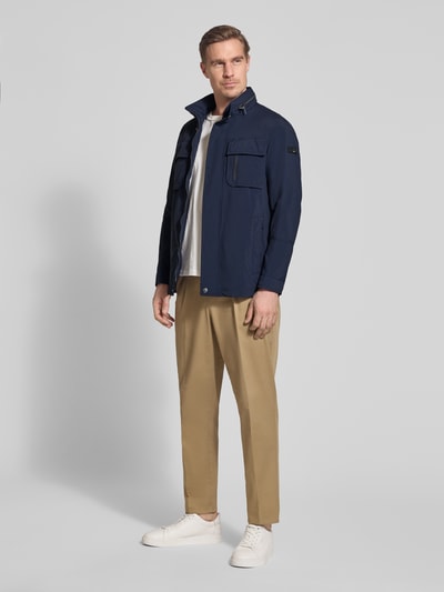 Męski model nosi Regular Fit Jacke mit Stehkragen od Bugatti w kolorze granatowym z kieszeniami na piersi, aplikacja z logo, krój regular fit, stójka, chowany kaptur, długość tyłu w rozmiarze 50: 72 cm, szerokość ramion: 46 cm, szerokość klatki piersiowej: 52 cm, długość rękawów: 68 cm, widok całej sylwetki z przodu.