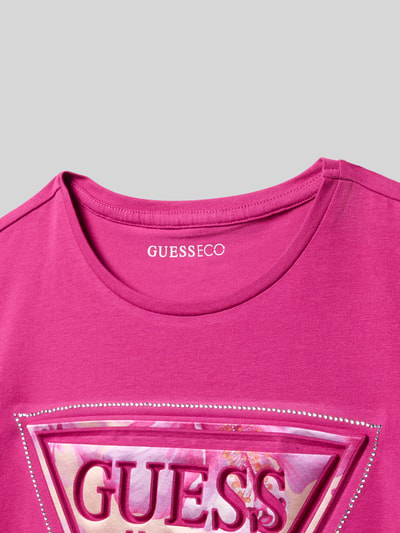 Produktfoto, T-Shirt mit Strasssteinbesatz und Logo-Print von Guess, różowy bawełniany materiał, nadruk logo, ozdobne dżety, okrągły dekolt, dziecięca kolekcja, lekko rozciągliwy materiał, wydłużony tył, luźny krój