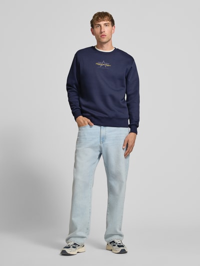 Männermodel trägt Regular Fit Sweatshirt aus Baumwoll-Mix mit Label-Detail von Alpha Industries in Dunkelblau, Ganzkörperansicht von vorne. Rückenlänge bei Größe M 67 cm, Ärmellänge 63 cm, elastische Abschlüsse