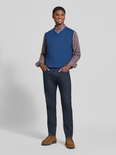 Männermodel trägt Regular Fit Pullunder aus reiner Baumwolle mit Logo-Stickerei von Fynch-Hatton in Blau über Karohemd, dunkle Jeans, Ganzkörperansicht von vorne. Rückenlänge bei Größe M 72 cm, V-Ausschnitt, gerippte Bündchen, ärmellos.