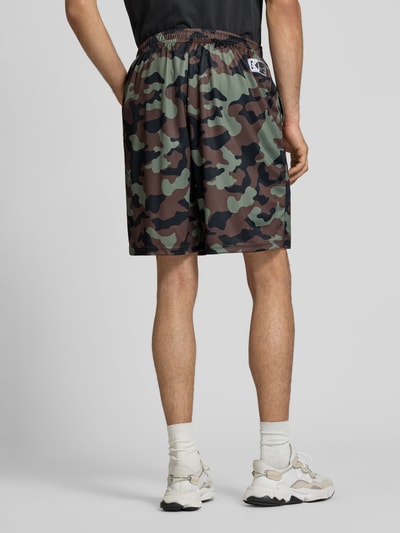 Männermodel trägt Regular Fit Shorts mit elastischem Bund von KARL KANI, Camouflage in Grün-Braun, Strukturmuster, Logo-Stitching, Ganzkörperansicht Rückansicht. Bundweite 38 cm, Innenbeinlänge 22 cm, Größe M.