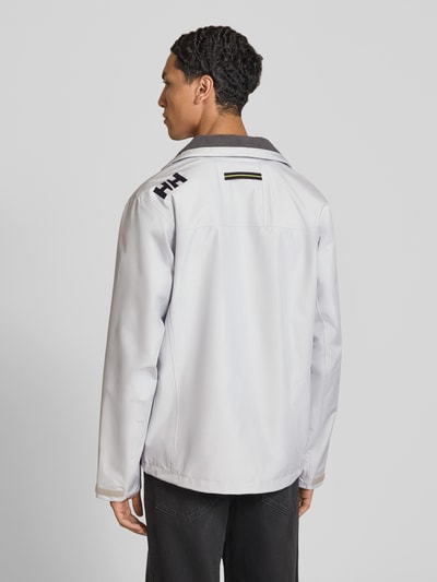 Männermodel trägt Regular Fit Jacke mit Stehkragen Modell 'Crew' von Helly Hansen in Weiß, Rückenansicht, Kontext: Stehkragen, Logo-Print, Napoleontasche, Reißverschlusstaschen, Reißverschluss, Rückenlänge 73 cm, Schulterbreite 48 cm, Brustweite 56 cm, Ärmellänge 68 cm. Ganzkörperansicht Rückansicht