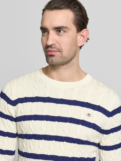 Männermodel trägt Slim Fit Pullover in Strick-Optik von Gant, cremefarben mit blauen Streifen, Logo-Stitching, Nahaufnahme Oberkörper.