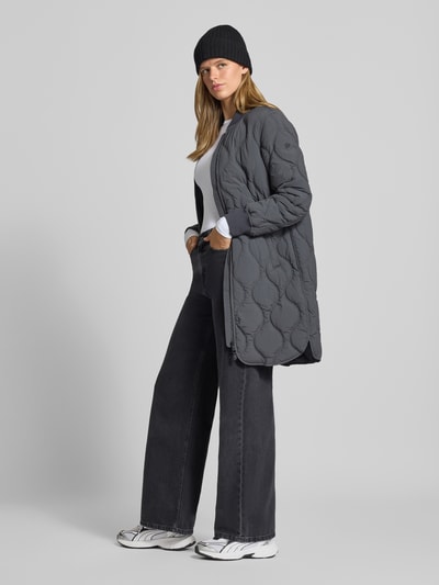 Frauenmodel trägt Steppjacke mit Stehkragen und Eingrifftaschen von Superdry, dunkelgrau, kombiniert zu weiter Jeans, Regular Fit, Ganzkörperansicht von vorne. Rückenlänge 91 cm, Schulterbreite 38 cm, Brustweite 50 cm, Ärmellänge 63 cm.