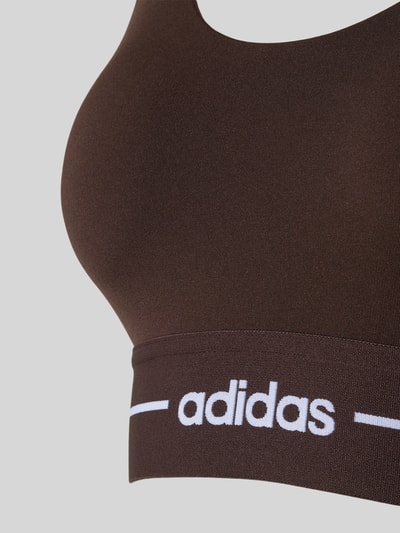 Frauenmodel trägt BH mit elastischem Logo-Bund von adidas, braun, körpernah, elastischer Bund, Material-Mix, Nahaufnahme Oberkörper.
