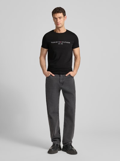 Männermodel trägt T-Shirt aus Bio-Baumwolle von Tommy Hilfiger in Schwarz mit Logo-Stickerei, gerader Schnitt, Ganzkörperansicht von vorne.