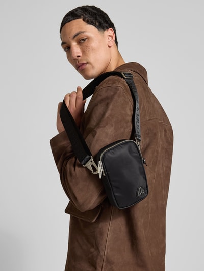 Männermodel trägt Crossbody-Bag mit Logo-Detail von Moose Knuckles in Schwarz mit Reißverschlüssen, Nahaufnahme Oberkörper. Die Tasche zeigt abnehmbaren Tragehenkel, Zweiwege-Reißverschluss und verstellbaren Trageriemen, Maße 11 x 18 x 4 cm.