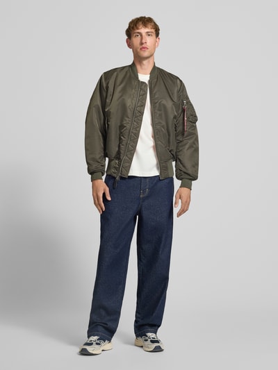 Männermodel trägt Bomberjacke mit Pattentaschen und Label-Applikation von Alpha Industries in Oliv, kombiniert mit Jeans; Ganzkörperansicht von vorne. Regular Fit, Stehkragen, Reißverschluss, sichtbar strukturierte Oberfläche. Rückenlänge 69 cm, Schulterbreite 44 cm, Brustweite 54 cm, Ärmellänge 64 cm.