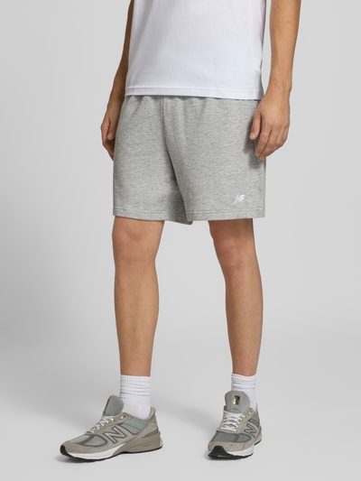 Männermodel trägt graue Sweatshorts aus Baumwoll-Mix Modell 'FRENCH' von New Balance, elastischer Bund, Logo, Nahaufnahme Beinbereich, Ganzkörperansicht von vorne.