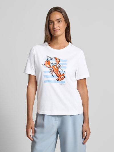 Modelka nosi T-Shirt mit Motiv-Print und Pailletten von Marc Cain, biała bawełna, krój regular fit, okrągły dekolt, motyw z cekinami, długość tyłu w rozmiarze 36: 56 cm, widok całej sylwetki z przodu.
