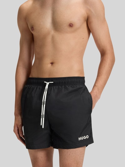 Männermodel trägt Badeshorts mit Label Print Modell 'HAITI' von HUGO, unifarben schwarz, seitliche Eingrifftaschen, Nahaufnahme Oberkörper.