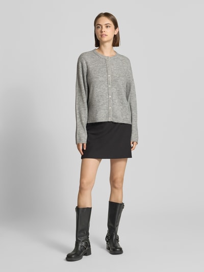 Frauenmodel trägt Loose Fit Strickjacke mit Rundhalsausschnitt Modell 'SIMONI' von ONLY in Grau, kombiniert mit schwarzem Minirock und schwarzen Stiefeln, Ganzkörperansicht von vorne, Damen Strickjacke von ONLY.