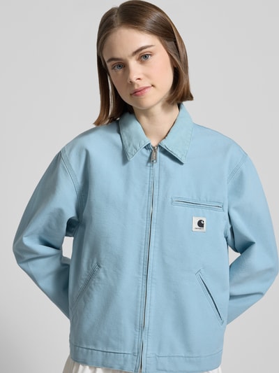 Frauenmodel trägt Jacke mit Label-Detail von Carhartt Work In Progress in Hellblau, Reine Baumwolle, Relaxed-Fit, Umlegekragen, Reißverschluss, Eingrifftaschen, Ganzkörperansicht von vorne. Rückenlänge bei Größe S: 58 cm, Schulterbreite: 47 cm, Brustweite: 54 cm, Ärmellänge: 60 cm.