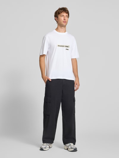 Mężczyznę model prezentuje biały t-shirt Regular Fit z czarno-żółtym nadrukiem, model 'Naxter' od HUGO, Męski t-shirt, zdjęcie całej sylwetki z przodu.