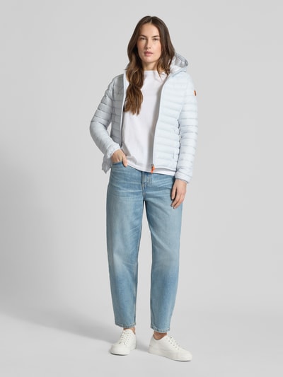 Frauenmodel trägt Slim Fit Steppjacke im Zweiwegereißverschluss Modell 'Daisy' von SAVE THE DUCK, helle Farbe, Stepp-Optik, Kapuze, Eingrifftaschen, Ganzkörperansicht von vorne.