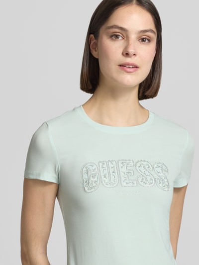 Frauenmodel trägt Slim Fit T-Shirt mit Strasssteinbesatz von Guess in Mint, elastischer Baumwoll-Mix, Rundhalsausschnitt, Logo-Detail, Nahaufnahme Oberkörper.