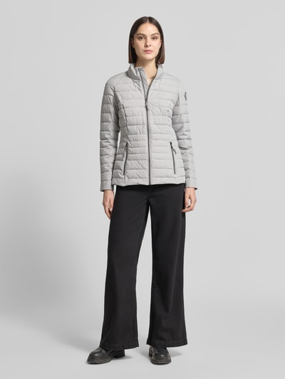 Modelka nosi jasnoszarą kurtkę pikowaną z zamkiem błyskawicznym i kieszeniami zapinanymi na zamek błyskawiczny, Stehkragen, krój slim fit, model 'GS 111' od G.I.G.A. DX, długość tyłu w rozmiarze 36: 67 cm, szerokość ramion w rozmiarze 36: 38 cm, szerokość klatki piersiowej w rozmiarze 36: 49 cm, długość rękawów w rozmiarze 36: 65 cm, widok całej sylwetki z przodu.
