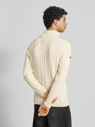 Männermodel trägt Strickpullover aus reiner Baumwolle von Superdry mit Zopfmuster, Troyer-Kragen, Label-Detail, Gerippte Abschlüsse, Rückenlänge 70 cm, Ärmellänge 71 cm, enge Passform, cremefarben, Ganzkörperansicht Rückansicht.