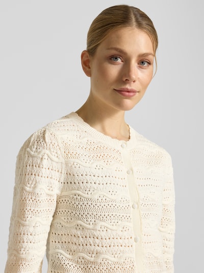 Modelka kobieca nosi Strickjacke mit Rundhalsausschnitt Modell 'GANACH' od SUNCOO PARIS, kolor ecru, czysta bawełna, krój regular fit, guziki, długość tyłu 52 cm, długość rękawa 34 cm; zbliżenie na górną część ciała.