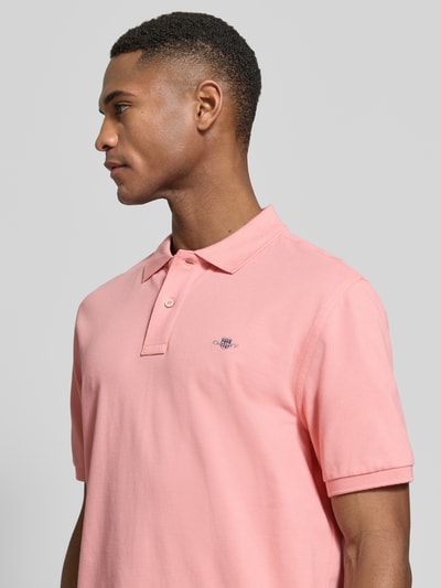 Męski model nosi Slim Fit Poloshirt mit Logo-Stitching od Gant z bawełny, krój slim fit, kołnierzyk polo, krótki rękaw, różowy, wyhaftowane logo, widok z boku, zbliżenie na górną część ciała