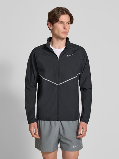 Männermodel trägt Regular Fit Jacke mit Stehkragen Modell 'Miler' von NIKE TRAINING in Schwarz mit Logo-Print und Reißverschluss, Ganzkörperansicht von vorne. Rückenlänge bei Größe M: 67 cm, Ärmellänge bei Größe M: 64 cm.