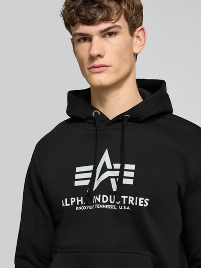 Männermodel trägt Hoodie mit Label-Print von Alpha Industries in Schwarz, Baumwollmischung, Unifarbenes-Design, Nahaufnahme Oberkörper.