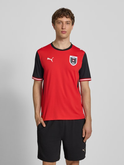 Männermodel trägt Österreich Heimtrikot 2026 von PUMA, rotes Sportshirt mit Logo-Print, Patch, Rundhalsausschnitt, Gerader-Schnitt, Ganzkörperansicht von vorne. Rückenlänge bei Größe M: 72 cm. Modell von PUMA PERFORMANCE.