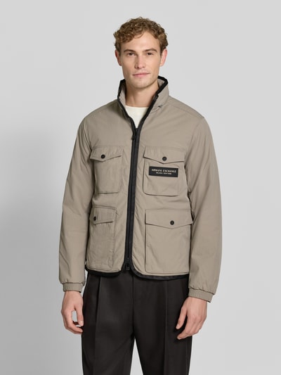 Männermodel trägt Regular Fit Jacke mit aufgesetzten Taschen Modell 'BLOUSON' von Armani Exchange in Beige mit Logo-Patch, Ganzkörperansicht von vorne.