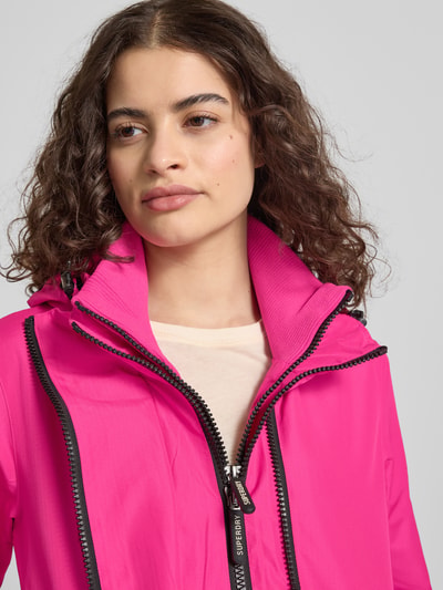 Frauenmodel trägt Windbreaker mit Zweiwege-Reißverschluss und Logo-Stitching von Superdry in Pink, Kapuze, Stehkragen, Nahaufnahme Oberkörper. Rückenlänge 63 cm, Schulterbreite 42 cm, Brustweite 37 cm, Ärmellänge 67 cm, Gerader-Schnitt, Seitliche Reißverschlusstaschen.