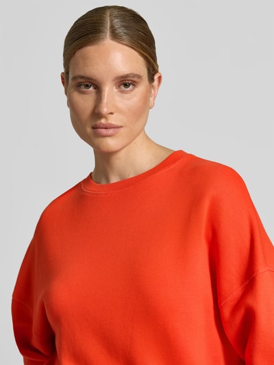 Vrouwenmodel draagt Oversized Sweatshirt aus Baumwoll-Mix van Juvia in feloranje, zachte textuur, ronde hals, Mix van katoen en modal, oversized, verlaagde schouder, elastische boorden, ruglengte maat S 54 cm, Nahaufnahme bovenlichaam.