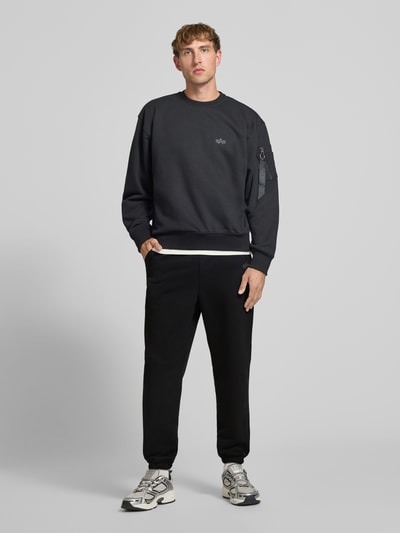 Männermodel trägt Sweatpants mit Label-Print von Alpha Industries in Schwarz, lockere Passform, Ganzkörperansicht von vorne, Baumwoll-Mix, Elastikbund, seitliche Eingrifftaschen, Innenbeinlänge 44 cm bei Größe M.