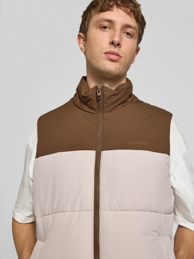 Männermodel trägt Weste mit Stehkragen Modell 'RAZE' von JACK & JONES in Braun und Beige, glatte Textur, Nahaufnahme Oberkörper, Regular Fit, Label-Print, Reißverschluss, Eingrifftaschen, KI-modifiziertes Bild.