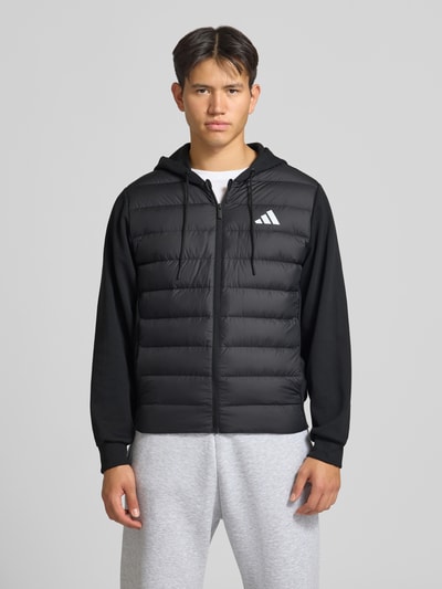 Männermodel trägt Regular Fit Steppjacke mit Kapuzenkragen von adidas Sportswear in Schwarz, glatte und gesteppte Textur, Ganzkörperansicht von vorne.