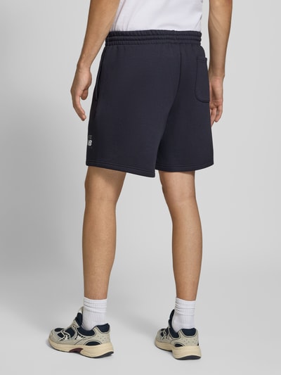 Männermodel trägt dunkelblaue Sweatshorts aus Baumwoll-Mix von New Balance mit elastischem Bund, Label-Print, Gesäßtasche, gerade Passform, Ganzkörperansicht Rückansicht.