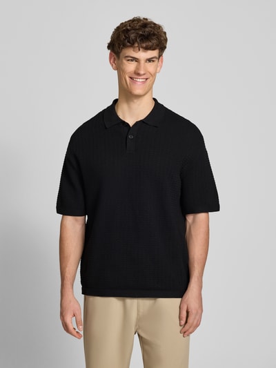 Männermodel trägt Relaxed Fit Poloshirt Modell 'FERDINAND' von Redefined Rebel aus Reine Baumwolle, Strickshirt, Knopfleiste, Rückenlänge 70 cm bei Größe M, schwarze Struktur, beige Hose, Ganzkörperansicht von vorne.