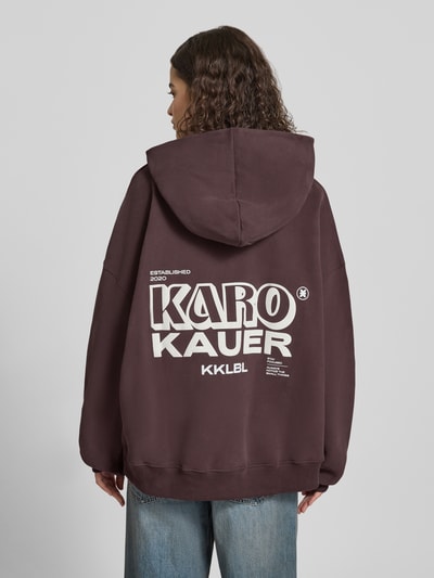 Frauenmodel trägt Oversized Hoodie mit Label-Prints und Känguru-Tasche von Karo Kauer in Dunkelbraun, reine Baumwolle, Ganzkörperansicht Rückansicht.