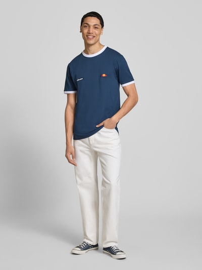 Męski model nosi T-Shirt z logo i naszywką label model 'RINGETTO' od Ellesse, bawełna, krój regular fit, widok całej sylwetki z przodu.