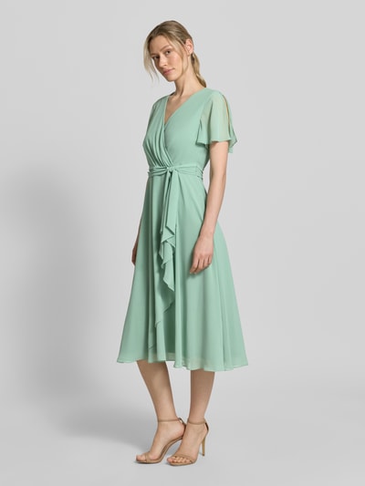 Vrouwenmodel draagt knielang cocktailjurk met strikceintuur van Christian Berg Cocktail, mintgroene chiffon, getailleerd model, wikkellook, volledige lichaamsvoorzijde.