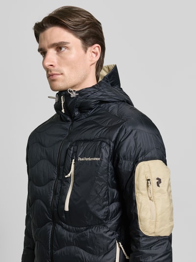 Männermodel trägt Regular Fit Jacke in Stepp-Optik mit Logo-Stitching von PEAK PERFORMANCE, Schwarz-Beige, Napoleontasche, Nahaufnahme Oberkörper.