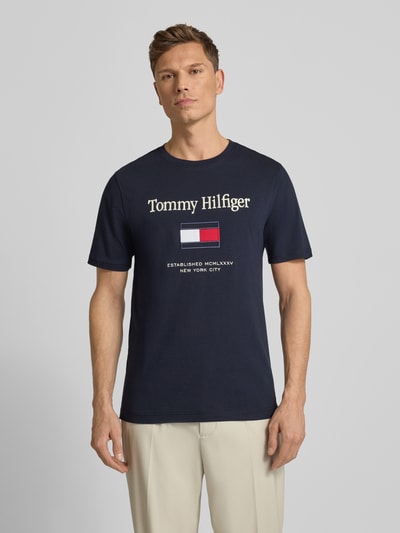 Męski model nosi Regular Fit T-Shirt aus reiner Baumwolle von Tommy Hilfiger, granatowa bawełna, nadruk z logo, krótki rękaw, widok z przodu, ujęcie całej sylwetki.