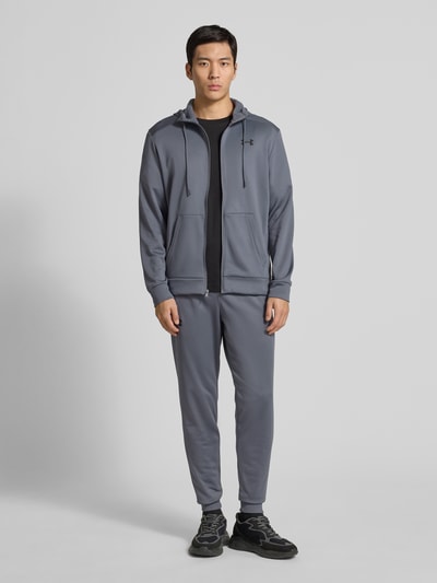 Männermodel trägt Sweatjacke mit Kapuze von Under Armour in Grau, Lockerer-Schnitt, Tunnelzug, Reißverschluss, Label-Detail, Eingrifftaschen, gerippte Abschlüsse, Rückenlänge 69 cm, Ärmellänge 66 cm, Ganzkörperansicht von vorne.