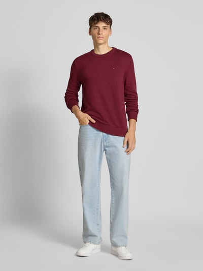Männermodel trägt Slim Fit Pullover aus reiner Baumwolle von Tommy Jeans in Uni-Bordeaux, Stricktextur, Rundhals, Langarm, Ganzkörperansicht von vorne.