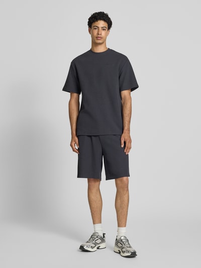 Männermodel trägt Relaxed Fit Shorts mit Strukturmuster von KARL KANI aus Baumwoll-Mix in Dunkelgrau, elastischer Bund, Ganzkörperansicht von vorne.