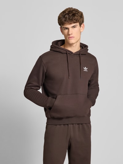 Męski model nosi Hoodie mit Kapuze od adidas Originals w kolorze brązowym, bawełniany miks, luźny krój, przód z kapturem, tunel, haftowane logo, kieszeń kangurka, ściągacze na zakończeniach, długość tyłu w rozm. M: 66 cm, długość rękawa: 66 cm, całkowity widok z przodu