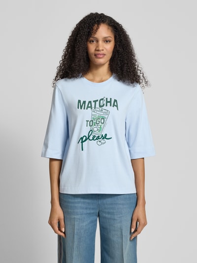 Modelka nosi Slim Fit T-Shirt mit Motiv-Print von Marc Cain, bawełna, krój slim fit, jasnoniebieski z nadrukiem, półrękaw, okrągły dekolt, długość tyłu 58 cm w rozm. 36, ujęcie całej sylwetki z przodu.
