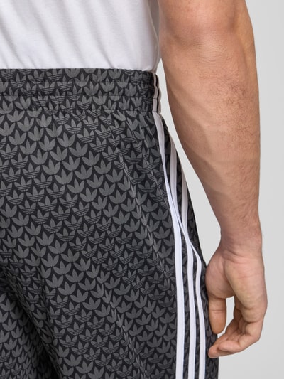 Männermodel trägt Trainingshose mit Logo-Muster von adidas Originals, Relaxed-Fit, Label-Stitching, elastischer Bund, Nahaufnahme Hüfte.